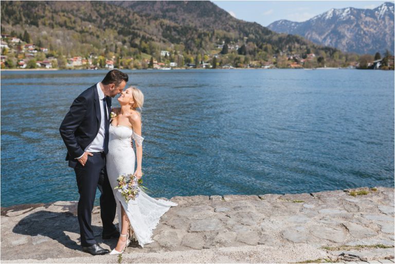 Brautpaar steht auf Steinweg vor dem Tegernsee und küsst sich