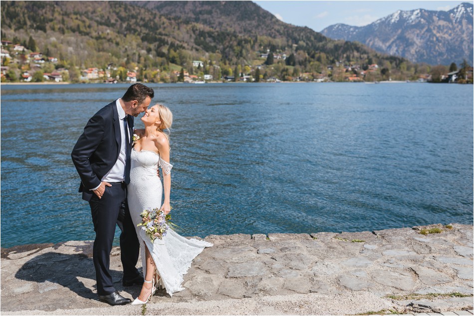 Brautpaar steht auf Steinweg vor dem Tegernsee und küsst sich