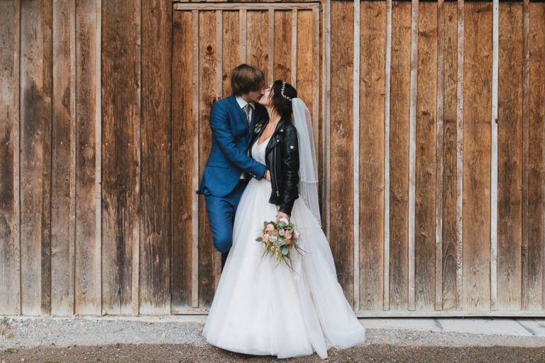 Brautpaar küsst sich vor Holzwand mit schwarzer Jeansjacke