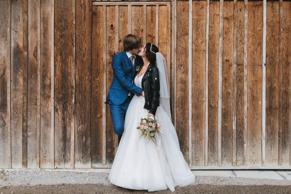 Brautpaar küsst sich vor Holzwand mit schwarzer Jeansjacke