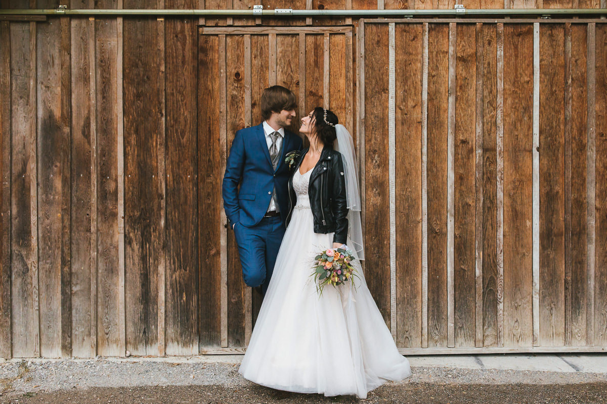 Brautpaar mit Leerjacke vor einer Scheunenwand bei ihrer Regenhochzeit